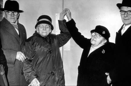 Photograph showing two elderly women dressed in coats and hats, looking towards the camera, while both holding a key up to a door; they are flanked on either side by a middle-aged man in overcoat, hat and glasses; the photograph has been identified as Opening of Pentecostal Church, Horden
