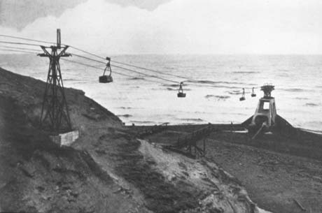 Photograph showing a pylon on a hillside with an aerial ropeway leading to a concrete structure at the edge of the sea; four containers are on the ropeway conveying coal waste to be disposed of in the sea; the photograph has been described as Aerial Flight, Horden