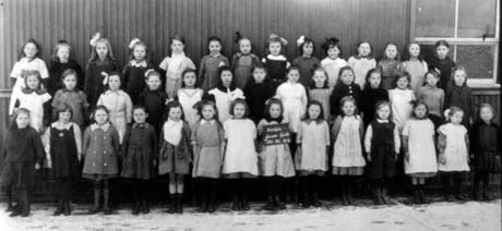 Photograph of forty two girls, aged approximately seven years, posed in three rows against the wall of a corrugated iron building; a girl on the front row is holding a notice reading: Horden Junior Girls Standard 3a 1919
