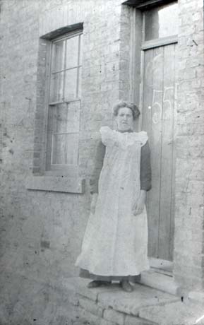Photograph of a woman standing on a step, above what appears to be a considerable drop, in front of the door and window of a brick house; the number 55 is written on the door in what appears to be chalk; the woman is wearing an ankle-length pinafore over a dark dress; the photograph has been described as Maher's Cottage and Housewife, Horden