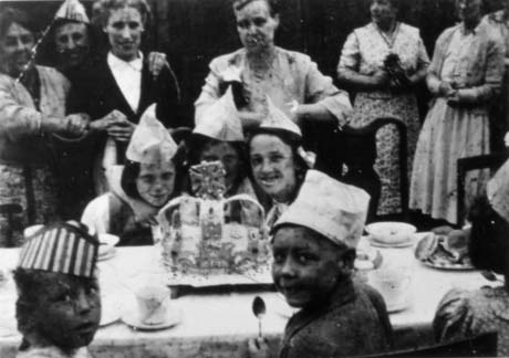 Photograph five children sitting at a table covered in a cloth and bearing crockery, a plate of buns, and a cake decorated with a crown; the children are wearing paper hats; behind the children on the far side of the table six women dressed in frocks and pinafores; the photograph has been identified as Morpeth Street Coronation Party, Horden