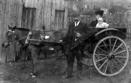 Photograph showing a horse harnessed to a two-wheeled trap in which a woman, wearing a dark hat and coat, and an infant are sitting; a man wearing a suit and cap and holding a pipe in his left hand is standing in front of the horse; behind the trap are wooden shed, a wooden fence; and beyond them, the backs of houses; they have been identified as Mr. and Mrs. Cooper with daughter, later Mrs. Robinson
