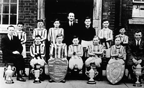 Photograph showing eleven boys, aged approximately thirteen years, wearing football strip, posed in two rows against the entrance of a brick building; they are accompanied by three men in suits; in front of the boys are four trophy cups and two shields; one of the shields reads: Easington Colliery School Football Club 1923