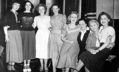 Photograph of six young women and two middle-aged women posed in a row inside a building; they are wearing jumpers, skirts and dresses; one of the young women appears to be sitting on the lap of one of the middle-aged women; they have been identified as Heddy Chorus Girls, Horden