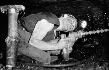 Photograph of a miner crouched double with a pit prop next to him indicating the shallowness of the seam in which he is working; he is holding an electric drill in his hand with a screw pointing at the wall of coal; the photograph has been identified as Electric Drill in Mine Shaft in Horden