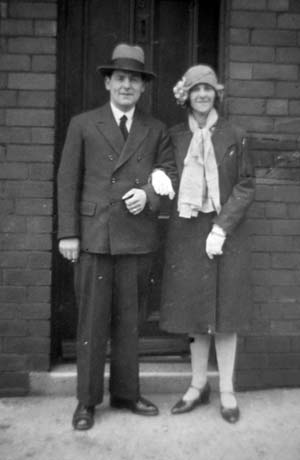 Photograph of a man, dressed in a double-breasted suit and tie and trilby hat, standing outside the doorway to a brick building, accompanied by a woman, dressed in light stockings, a dark coat, light gloves and a cloche hat with a decoration on its side; they have been identified as Mr. and Mrs. Vinsome of Horden