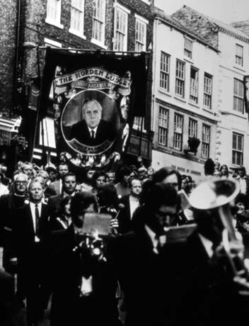 Photograph showing the Horden Lodge banner being carried in Sadler Street in Durham City during the Durham Miners' Gala; the portrait of Harold Wilson, Labour Prime Minister, 1964-1970 and 1974-1976, can be seen on the banner; men playing musical instruments are walking in front of the banner