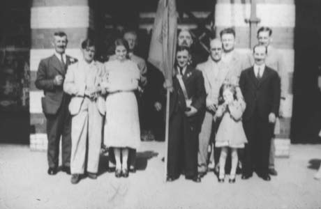 Photograph showing one woman in a frock, nine men in suits, and one small girl in a coat and frock, aged approximately seven years, standing with a flag, which cannot be identified, in front of the imposing doorway of a building; they have been identified as British Legion Members, Horden