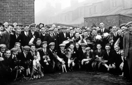Photograph of a crowd of men, wearing suits and caps and holding large vegetables, presumably grown for a show, posed with the front row kneeling and the two rows behind standing; behind the group are the backs of terraced houses; they have been identified as Buffs Members in Horden