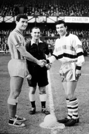 Photograph of two men in football strip shaking hands across a football in a football stadium with crowds in the stands; a referee is standing between the two men who have been identified as S. Anderson and C. Hurley
