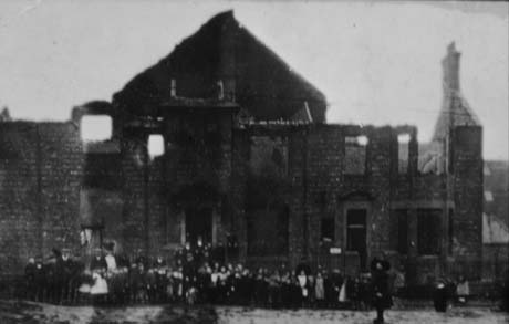 Photograph showing the indistinct outline of the ruined roof and walls of a large building with a crowd in front; the photograph has been described as Horden Big Club After The Fire