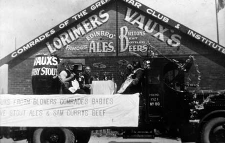 Photograph showing the side of a motor lorry parked in front of a single-storey wooden building with the following written on it: Comrades Of the Great War Club And Institute. Lorimers Famous Edinbro' Ales. Vaux's Britons Best Bottled Beers; the lorry is decorated as a float with a barrel on its back and a table with beer bottles on it, round which three men are sitting; on its side is a notice reading: Vaux Froth Blowers Comrades Babies Stout Ales & Sam Currys Beef; the photograph has been described as Bob Ridley Beer Float, Horden