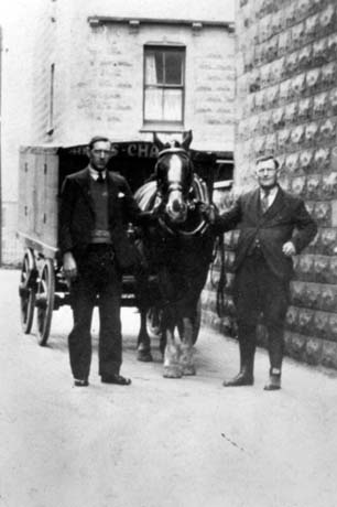 Photograph showing a horse facing the camera harnessed to a cart with a covered container on it; a man dressed in a jacket, breeches and leggings is standing on the right of the horse at its head with his back to the stone wall of a house; on the other side of the horse's head is a man in a suit and tie; behind the group is the end of a stone house; the photograph has been described as Charlie Williams in Hardwick Street, Horden