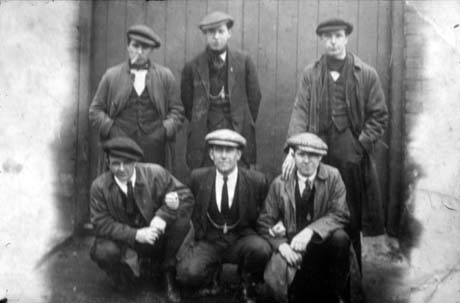 Photograph showing three men standing, and three men squatting, in front of a large wooden door; they are wearing suits, waistcoats, raincoats, and caps; they have been identified as Group of Miners, Horden