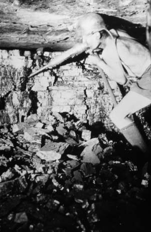 Photograph showing a miner dressed in socks, shorts and helmet, using a drill on a wall of coal; the ceiling immediately above the miner's head and pieces of coal on the floor can be seen; the photograph ahs been described as Nig Nog, Horden