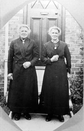 Photograph of two elderly women standing side by side with a doorway and steps behind them; they are wearing dark ankle-length dresses; they have been described as Twin Sisters, Horden and they have been given the names Mrs. Lang on the left and Mrs. Dawe on the right