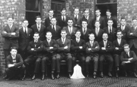 Photograph showing twenty five men, in suits and ties, posed in three rows against a brick building with sash windows; there is a cardboard bell in front of them; they have been identified as Bell Club Snooker Team, Horden