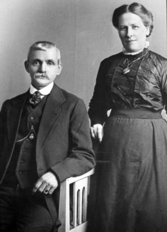 Photograph of a man wearing a suit, tie, waistcoat and watch chain, sitting in a wooden chair with arms; next to him a woman, wearing a dark blouse, dark skirt, and locket is standing next to him; they have been identified as Thomas and Isabella Vinsome, Horden