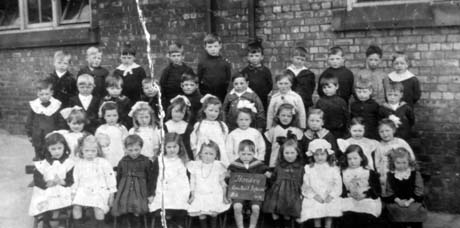 Photograph of forty one children, aged approximately six years, posed in four rows against the wall of a brick building; a child on the front row is holding a notice reading: Horden Council Infants 2D 1919