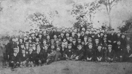 Photograph of approximately one hundred and twenty men posed with trees behind them; they are all wearing suits and caps; they have been identified as Miners on Strike Fighting Against 10-Hour Work Days, Horden