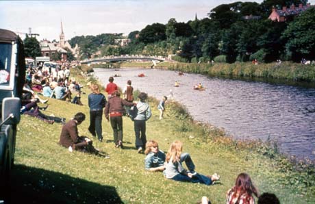Photograph showing the River Wear, running through Durham City, on the right of the photograph with young people sitting on the river bank on the left of the photograph; all of the young people on the river bank have their backs to the camera; five canoes can be seen in the distance on the river; a bridge and the spire of a church can be seen in the distance; the photograph has been identified as having been taken during the Miners' Gala