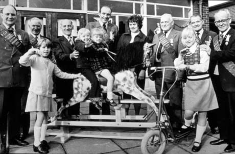 Photograph showing seven men, wearing suits and sashes across their chests, standing in a semi circle round two children sitting on a rocking horse and a child standing on a scooter; another child is standing with her hand on the rocking horse; a woman is watching the children; behind the group is a building largely of glass; the photograph has been identified as Buffs Presentation For Autistic Children, Horden