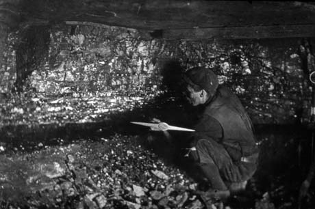Photograph of a miner, wearing leather jerkin and trousers, crouching in front of a wall of coal with a pick in his hand; he has been identified as being in Horden