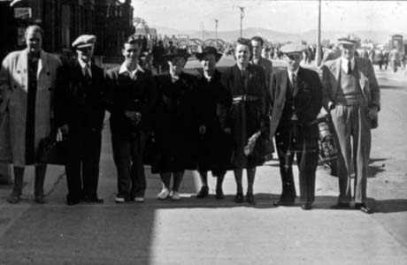 Photograph showing four men and four women, wearing overcoats and suits and hats, standing in a row across a pavement with crowds, and hills in the distance; they have been described as members of The Good Templars on an outing