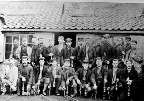 Phtograph of a group of twenty three men wearing work clothes and carrying miner's lamps posed in two rows outside a single-storey building with a tile roof; the men on the front row are also holding pointed metal objects with a hole in the middle; they have been identified as miners in Horden
