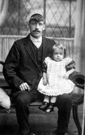 Photograph showing a man in a dark suit sitting on a chaise longue with a small girl aged, approximately two years, sitting on his knee; behind them is a window; the small girl is wearing a broderie anglaise pinafore over a tartan dress, and boots; they have been identified as Mr. Cooper and his daughter, Horden; Mr. Cooper has been identified as a worker at Nimmo's Brewery