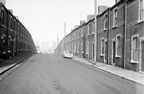 Photograph showing both sides of a road of terraced houses running away from the camera; at the end of the road two large towers and the winding gear of a colliery can be seen; the road has been identified as Colliery Street, Horden; two cars are parked on the road