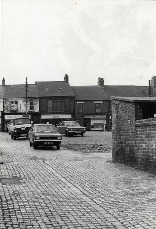 Photograph showing the facades of three shops across an open space, possibly the result of demolition; the name of the only shop that can be read is Elliot & Lee; three cars are parked in the open space, the registration of one of which is STM 60N; in the foreground is a cobbled surface and the wall of a garden and outhouse; they have been identified as being in Horden