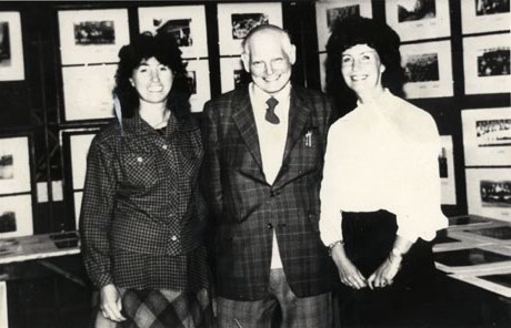 Photograph of an elderly man wearing a checked jacket and trousers with a woman in a checked skirt and checked blouse on the left of him and a woman in a light-coloured blouse and a dark skirt on the right of him; behind them are display boards with photographs of scenes on them; they have been identified as Ivy Summerson; Margaret Hunt; Ken Richardson and as being in Horden