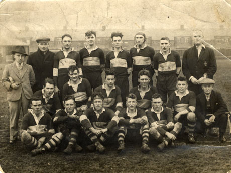 Photograph of sixteen men in rugby strip posed on a field with houses in the distance; they are accompanied by four other men in suits and blazers; they have been identified as being in Horden