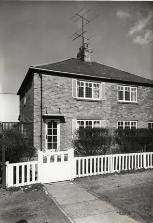 Photograph of the exterior of the front of a brick semi-detached house with one window on the ground and first floors and a glass door; a low fence is in front of the garden with a hedge behind; the house has been identified as being in Horden