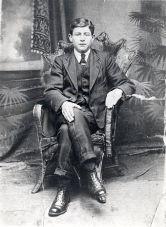 Photograph of a man wearing a suit, waistcoat, tie and boots, sitting in a chair in a photographer's studio; he has been identified as an inhabitant of Horden