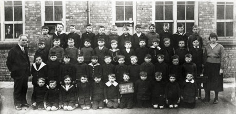 Photograph of a group of fifty boys, aged approximately seven years, posed in four rows in front of a brick building with large windows; a man is standing at the left and a woman at the right; a child on the front row is holding a notice reading: Horden Junior Boys Standard 2a 1921