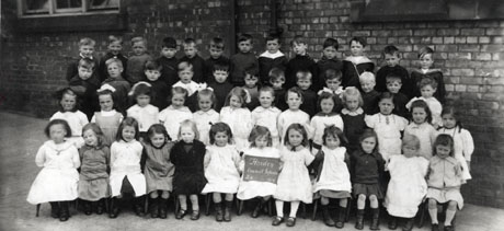Photograph of fifty one children, aged approximately six years, posed in four rows in front of a brick wall; a child on the front row is holding a notice reading: Horden Council Infants 2A 1919
