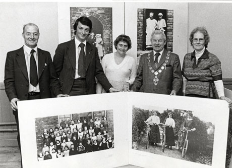 Photograph showing two men in suits, a woman wearing a light-coloured jumper, a man in a suit wearing a chain of office and a woman wearing a patterned jerkin, holding a large copy of two archive photographs; there are two archive photographs on the wall behind them; they have been identified as being in Horden