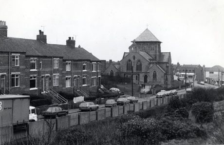 Photograph showing the exterior of east end of the church of St. Mary, Horden, and the front of a terrace of houses leading to the church; in front of the terrace is a road in which ten cars and a Co-op van are parked; in the foreground are a fence and rough vegetation