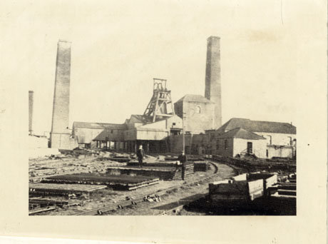 Photograph of the exterior of the surface buildings at Hutton Henry Colliery, showing two chimneys and the winding gear; in the foreground rail tracks can be seen with two boys standing on piles of material; to the right of the picture empty coal trucks can be seen; the photograph is described as Hutton Henry Upcast Shaft