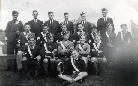 Photograph of a group of thirteen boys, members of the Boys' Brigade, wearing their uniforms and holding musical instruments; behind the boys, the six adult officers of the Brigade, also in uniform, are standing; the group is photographed in the open air, possibly at a camp