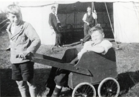 Photograph of a boy of approximately ten years of age pulling a cart in which another boy is sitting; behind the boys a marquee can be seen with two men standing laughing at the opening of the marquee; the photograph has been identified as Boy's Brigade- Sale of Work, Low Fields, Hutton Henry, 1936