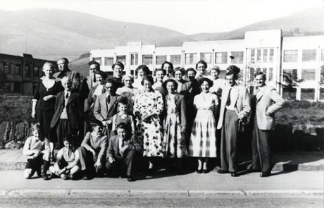 Photograph of twenty one men and women, dressed in suits and summer dresses, and five children, aged between approximately seven and twelve years, grouped in front of a large building, possibly a factory,with hills in the distance; the photograph has been identified as Hutton Henry Church Choir Outing, 1950s