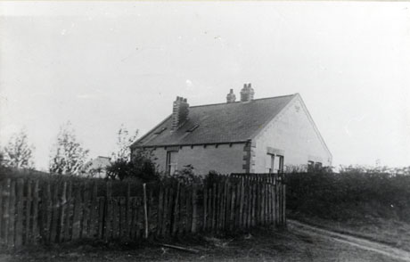 Photograph showing a fence in the foreground, beyond which the top storey and the slate roof of a building can be seen; the photograph has been identified as Woodbine Cottage, Hutton Henry