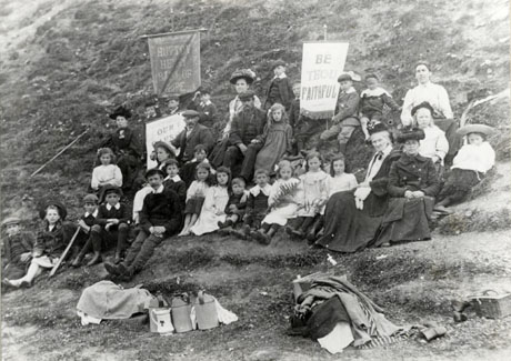 Photograph showing nine men and women and twenty four children sitting on a hillside accompanied by banners reading: Hutton Henry Band of Hope, Be Thou Faithful and Our Cause ...; in front of the group are piles of cloths, three flagons, a jug and a pic-nic basket; the photograph has been identified as Band of Hope at Hart