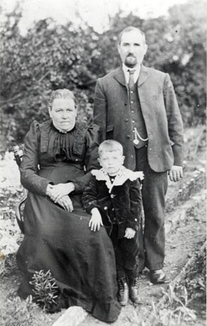 Photograph of a woman dressed in a dark long dress with pleats in the bodice, sitting on a chair with bushes in the background; man with a moustache and beard, wearing a suit, waistcoat and tie, is standing beside her; in front of them a small boy, aged approximately six years, wearing a velvet suit and fancy collar, is standing