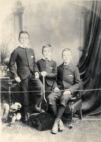 Photograph showing a boy, aged approximately ten years, wearing a suit and bow tie, leaning on a table on which there is a pot containing a plant; his left foot is resting on a footstool on which, also, a boy, aged approximately six years, wearing a suit and bow tie is resting a tennis racket, which he is holding in both hands; next to him is a boy, aged approximately twelve years and wearing a suit and tie, is sitting on a chair; the boys have three-quarter-length trousers; the photograph has been taken in a photographer's studio