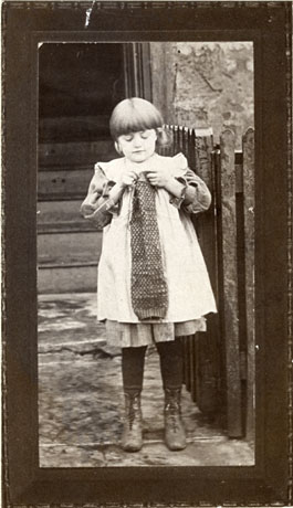 Photograph of a small girl, aged approximately ten years, wearing a dress, a pinafore, dark stockings and boots, standing knitting a long piece of knitting; behind her are a flight of steps leading into a building, a wall, and a fence