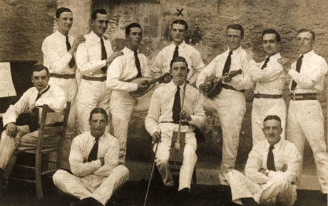 Photograph of seven young men, dressed in white trousers and shirts and dark ties and belts, standing against a wall;two of them are holding ukeleles; in front of them, three more young men, similarly dressed, are sitting; one is playing a stringed instrument; on the left of the picture is a young man sitting at a piano; they have been identified as The Cavaliers Band